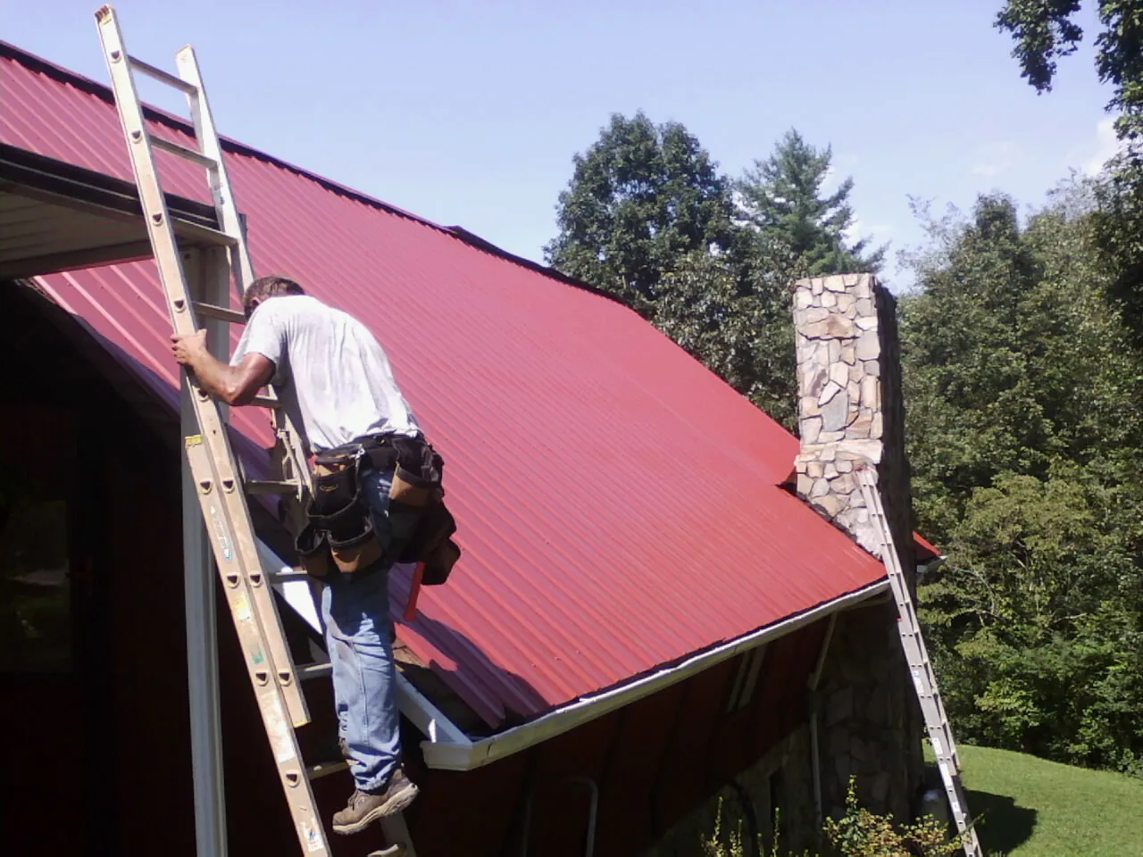 Roofer on a ladder beside a red metal roof and stone chimney — Soffit & Fascia Repair by Roofing Brothers in Greencreek